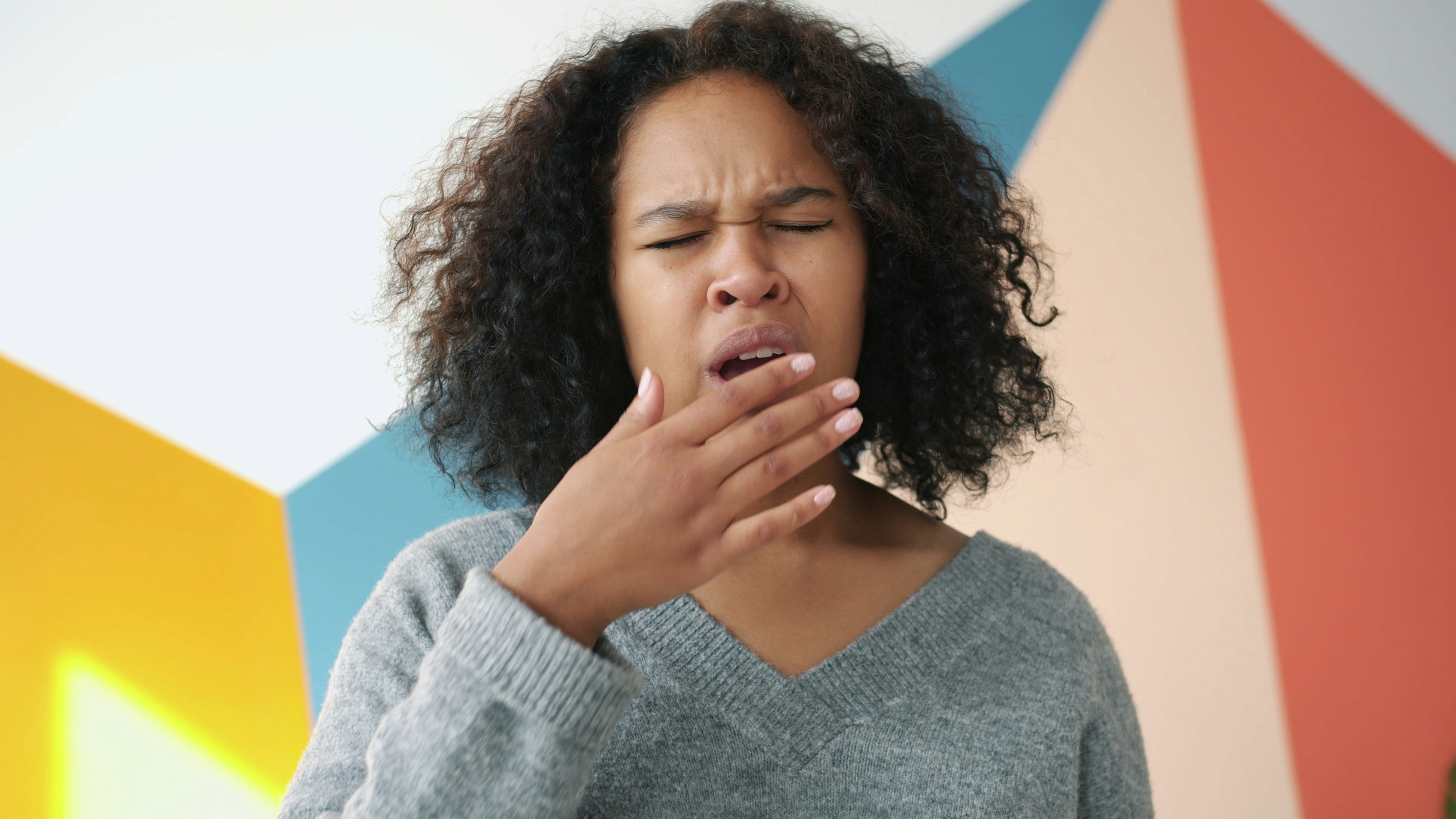 Portrait of sleepy Afro-American person pretty girl yawning looking at camera at home with colorful wall in background. Modern people and body language concept.